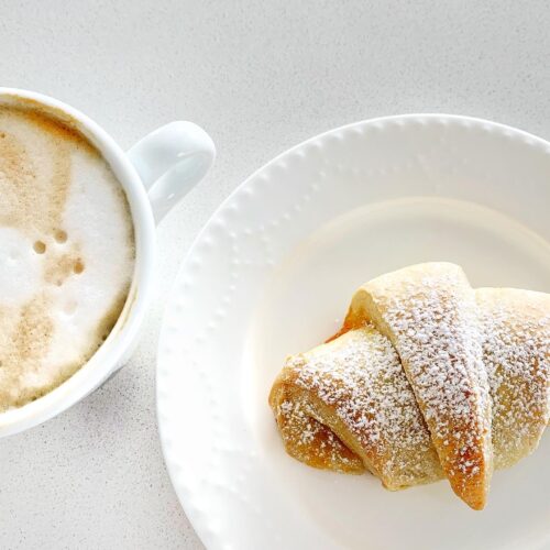 Homemade Italian cornetti served with cappuccino on a white plate, dusted with powdered sugar.
