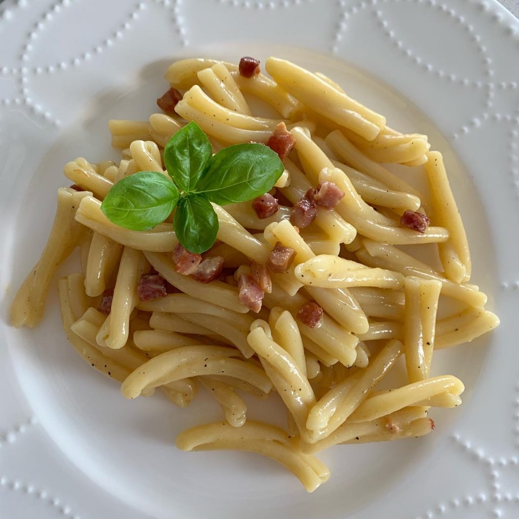Gemelli carbonara served on a white plate, topped with crispy pancetta and fresh basil.